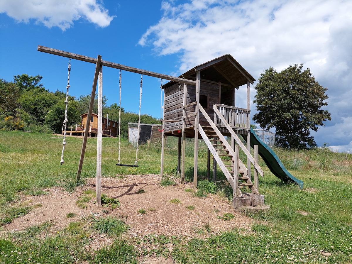 A wooden play structure features a raised platform with a roof, stairs, and a slide. Adjacent swings hang from a sturdy frame, inviting children for outdoor play. The surrounding grassy area includes low shrubbery and trees, under a partly cloudy blue sky.