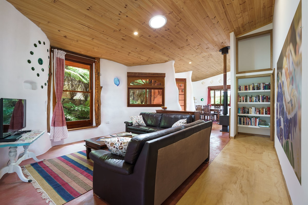 A cozy living area is highlighted with wooden beams and natural light from large windows. A dark leather sofa is positioned comfortably on a colorful rug. Bookshelves and a dining area can be seen in the background, complemented by vibrant wall art.