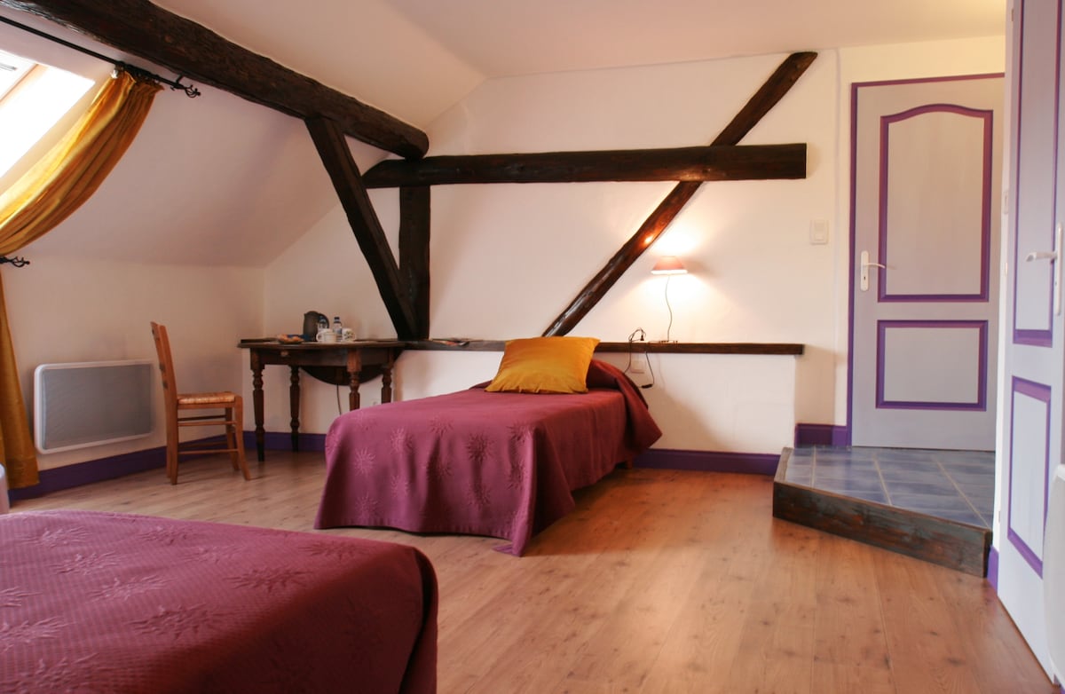 A room with a sloped ceiling features two single beds, each dressed in deep purple bedding. A wooden chair and a small desk are along one wall, with a decorative pillow placed on one bed. Exposed beams and a large window with light drapes add character.