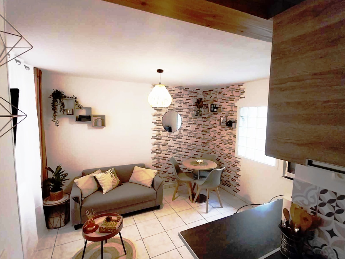A cozy living area is highlighted by a comfortable sofa adorned with decorative cushions. A round dining table with chairs is positioned nearby, complemented by a wall featuring a mix of brick and neutral tones. Natural light streams in through a window, enhancing the inviting space.