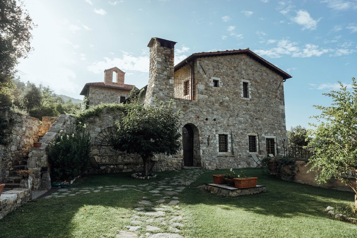 A rustic stone building features an arched entrance, surrounded by a manicured lawn. Pathways made of flat stones weave through the yard. A small garden with planters is situated in front, while trees and greenery provide shade in the background.