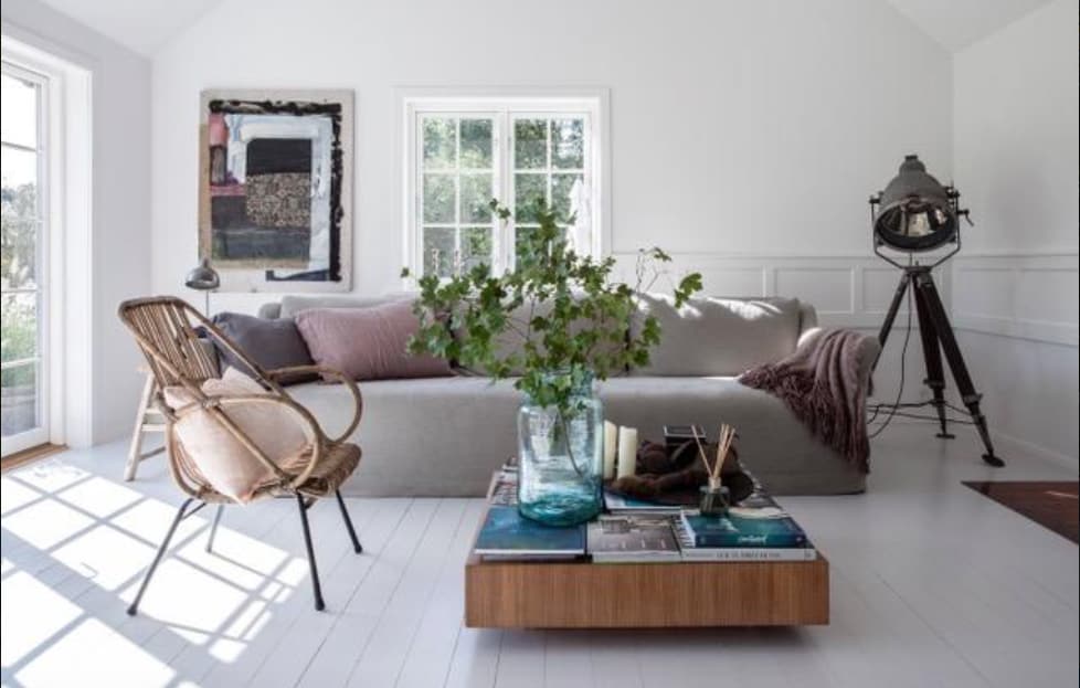 A light-filled living area features a spacious sofa adorned with neutral cushions. A wooden coffee table displays magazines, and a glass vase holds fresh greenery. Natural light enters through large windows, illuminating the room's simple yet stylish decor, complemented by a vintage-style floor lamp and a tripod light.
