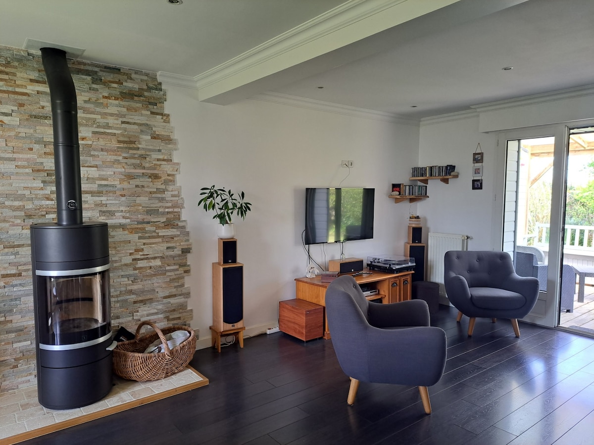 A spacious living area features a modern wood stove against a stone wall. A flat-screen TV is mounted on the wall, accompanied by a wooden cabinet and speakers. Two comfortable chairs in grey are positioned for relaxation, with natural light streaming in from nearby doors that lead to the terrace.
