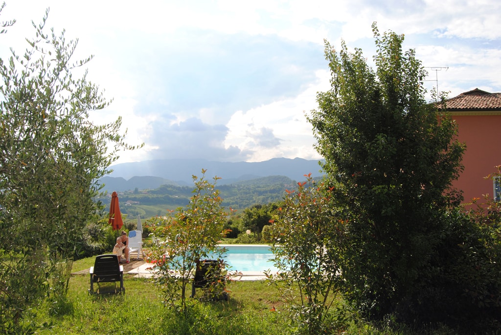 A serene outdoor area is depicted, featuring a swimming pool surrounded by lush greenery and colorful shrubs. A lounge chair and a sun umbrella are visible by the poolside, providing a relaxing space with panoramic views of the surrounding hills.