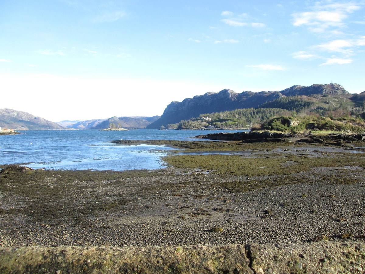 A calm waterfront view showcases a rocky shoreline leading into the tranquil waters of Loch Carron. The surrounding hills are visible in the distance, illustrating a natural landscape with a blend of earth tones and greenery under a clear blue sky.