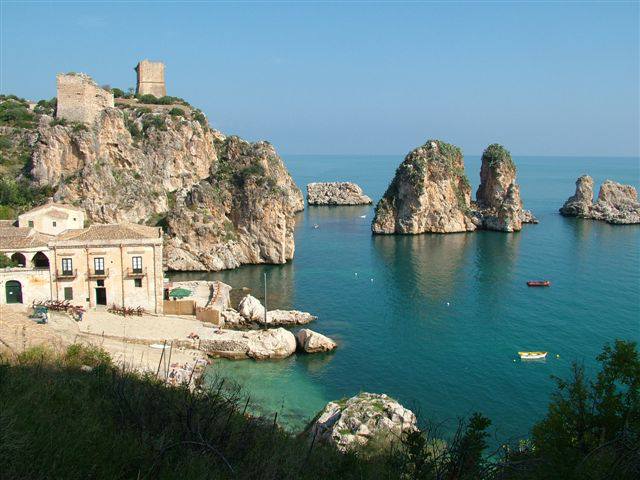 A tranquil coastal scene is showcased, featuring rocky formations rising from clear blue waters. In the background, a historic tower sits atop a hill, while a charming building is nestled along the shoreline, providing a peaceful ambiance by the sea.
