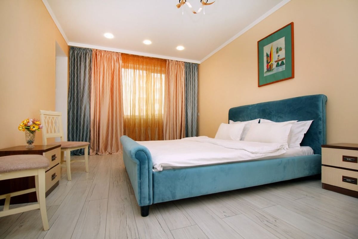 A spacious bedroom features a soft blue upholstered bed with white bedding. Natural light filters through sheer curtains, illuminating the room. Minimalist furniture, including a small table with a vase of flowers, complements the warm color scheme.