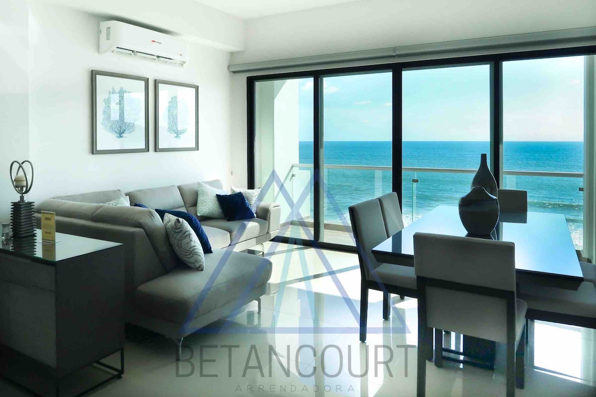 A well-lit living space features a comfortable sofa adorned with decorative pillows, positioned to face large glass doors that provide a panoramic view of the ocean. A dining table with chairs is present, complementing the modern decor and inviting ocean backdrop.