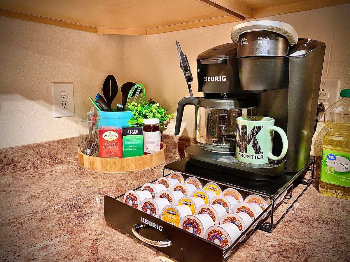 A coffee station is set up with a Keurig machine and a variety of coffee pods organized in a drawer. A decorative mug is placed nearby, accompanied by utensils and other supplies within a wooden tray. A small potted plant adds a touch of greenery to the countertop.