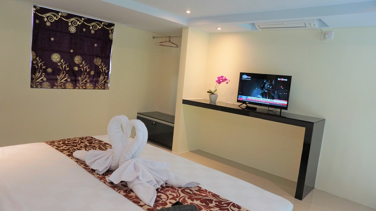 A well-appointed bedroom features a spacious bed adorned with neatly arranged towels in the shape of swans. A contemporary unit with a television is displayed against a light wall, and a small potted plant adds a touch of greenery beside the screen. Light filters in through a decorative window covering.