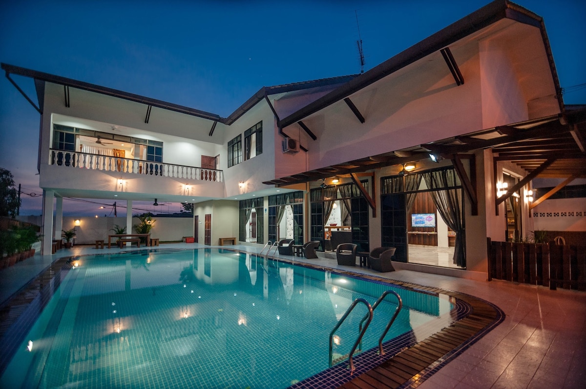 The exterior of the villa is illuminated at dusk, highlighting the spacious swimming pool that reflects soft lighting. A large terrace area is visible beside the pool, furnished with seating. The building features large windows and balconies that offer views of the surroundings.