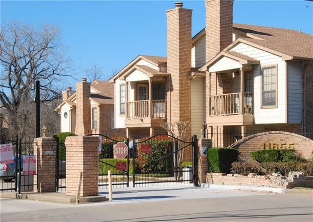 A gated community entrance is highlighted, featuring brick columns and a welcoming facade. Two buildings are visible, each with balconies and multiple windows. Green landscaping surrounds the area, adding to the neat and secure environment.