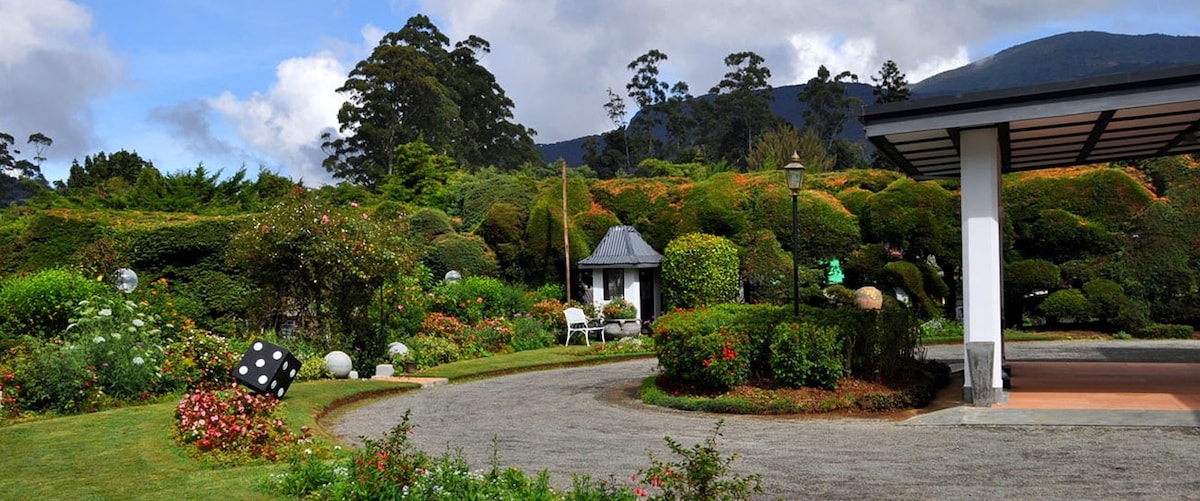 Lush gardens are featured, showcasing diverse greenery and colorful flowers. A charming gazebo is positioned amidst manicured hedges. Seating arrangements are visible alongside decorative stones, with rolling hills and cloudy skies in the background, creating a peaceful outdoor environment.