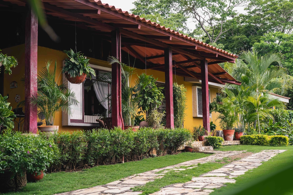 The exterior of the chalet features a welcoming porch with vibrant red columns and terracotta roof tiles. Lush greenery and potted plants line the pathway, which leads to a cozy entrance. Natural light filters through the large windows, enhancing the atmosphere of tranquility.