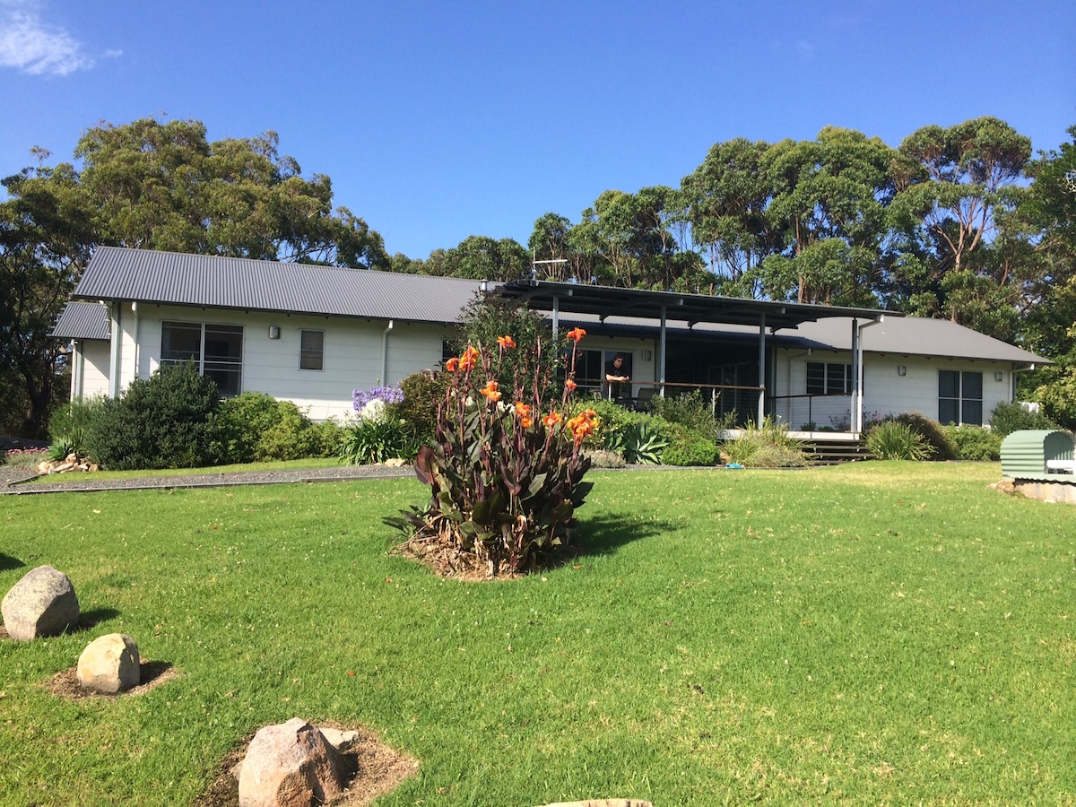 A modern home is set amidst lush greenery, featuring a spacious outdoor area. Colorful flowers are arranged in a garden bed in the foreground, while the house has large windows and a covered deck that invites relaxation. A clear blue sky complements the tranquil scene.