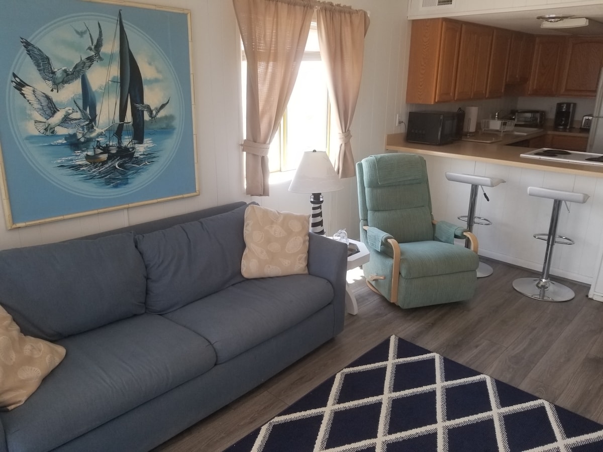 A cozy living area includes a blue sofa adorned with decorative pillows, positioned next to a light fixture and a green recliner. Natural light filters through the window, accentuating the adjacent kitchen's cabinetry and breakfast counter with stools. A geometric rug adds texture to the space.