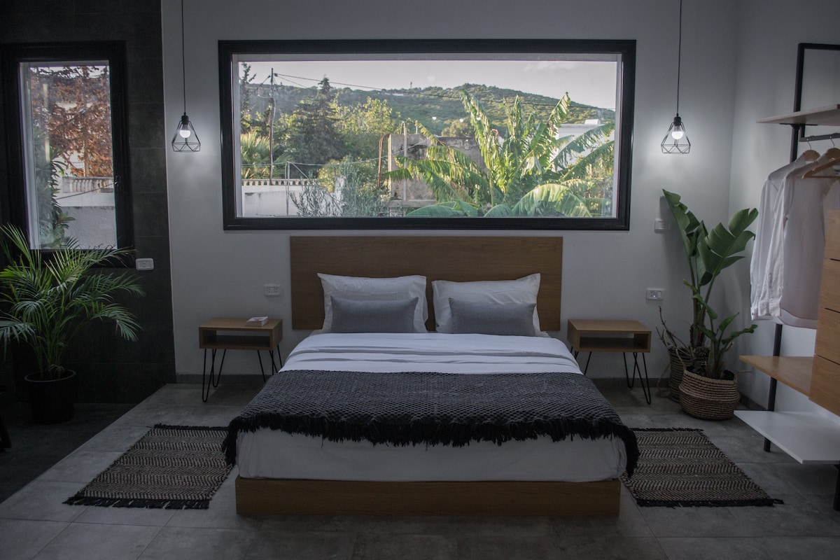 A modern bedroom features a spacious bed with neutral bedding, complemented by two bedside tables. Large windows frame views of greenery outside. Soft light emanates from hanging pendant lights, and a decorative rug adds warmth to the concrete floor, enhancing the serene ambiance.