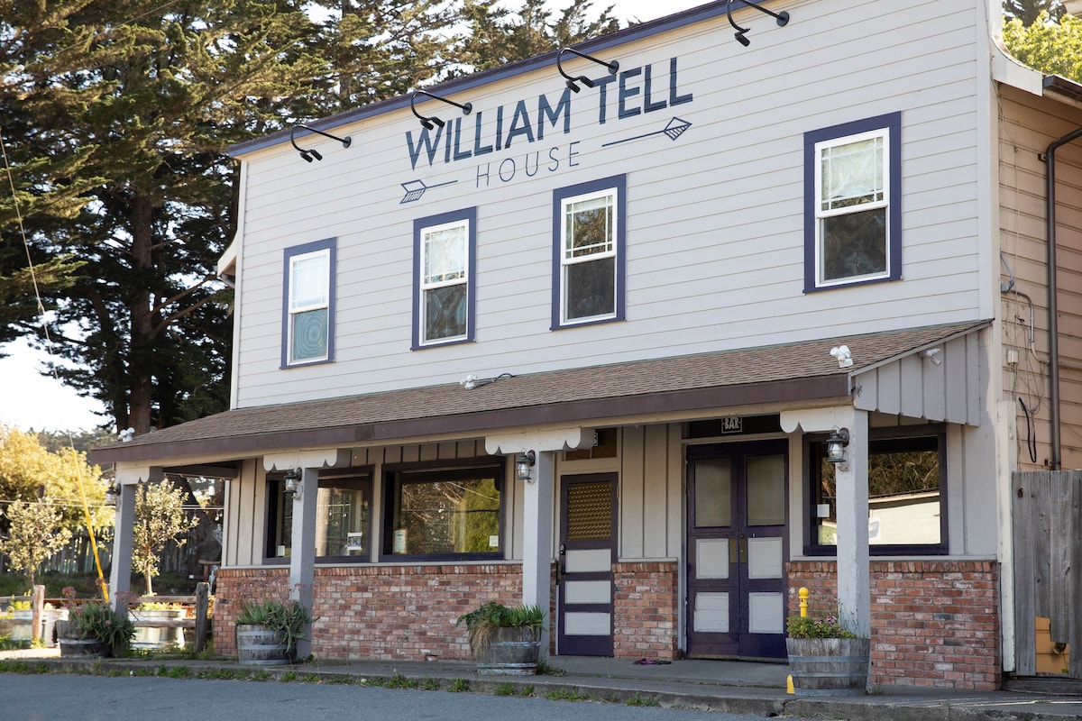 A two-story building features a welcoming façade with the name 'William Tell House' prominently displayed. Large windows are positioned above a covered entrance, and brick accents complement the exterior. Surrounding greenery enhances the charming curb appeal of this coastal establishment.