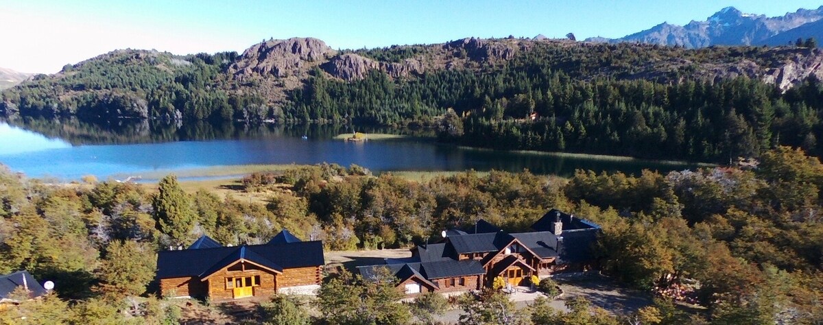 A panoramic view captures a lodge nestled among lush greenery with a serene lake in the foreground, reflecting the surrounding mountains. The architecture features traditional wooden elements, blending with the natural landscape, creating a sense of harmony with the Patagonia environment.