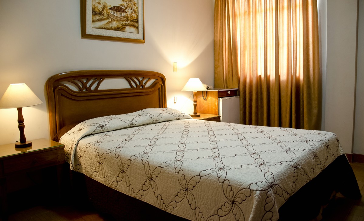 A spacious bedroom is illuminated by warm lighting from two bedside lamps. A patterned quilt covers the inviting bed, complemented by dark curtains at the window. Decorative artwork hangs above the headboard, while a small refrigerator is positioned near the wall.