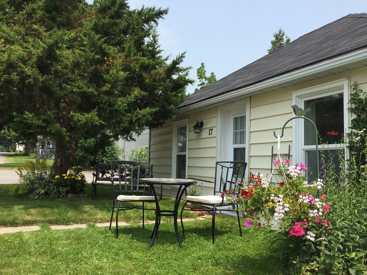 A charming outdoor seating area features a small round table with two chairs, nestled beside a vibrant flower bed. A large tree provides shade, and the cottage's entrance is framed by a welcoming garden, enhancing the calm neighborhood setting.