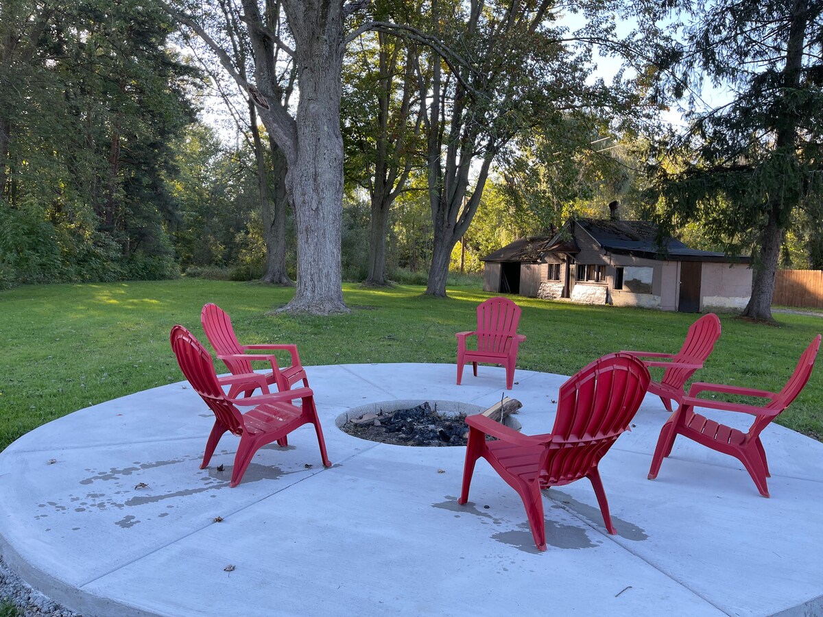 A circular concrete patio is surrounded by five red Adirondack chairs, providing a cozy gathering spot around a fire pit. Lush greenery and large trees create a serene outdoor atmosphere, with a rustic building visible in the background.
