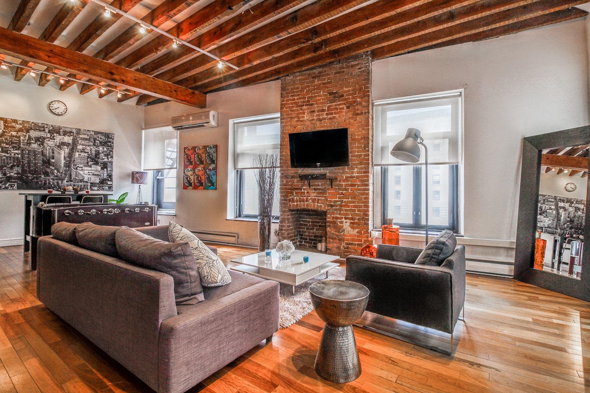 A spacious living area showcases a mix of modern and classic elements with exposed wooden beams and a rustic brick wall. Comfortable seating is arranged around a glass coffee table, complemented by a large mirror and vibrant artwork. Natural light filters through large windows.
