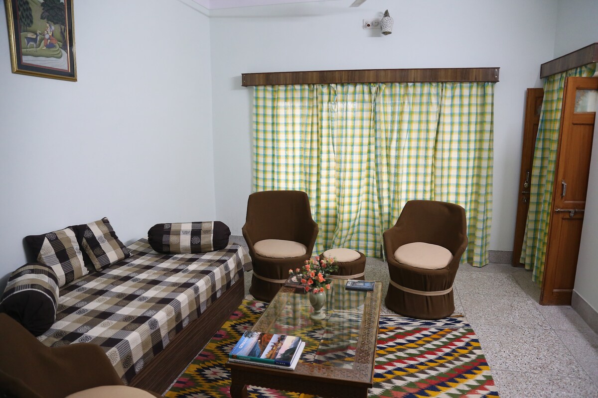 A comfortable seating area is presented, featuring two upholstered chairs and a daybed, adorned with a checkered pattern. A coffee table sits centrally, complemented by a small floral arrangement. Light filters through the green and yellow checked curtains, enhancing the room's calm atmosphere.