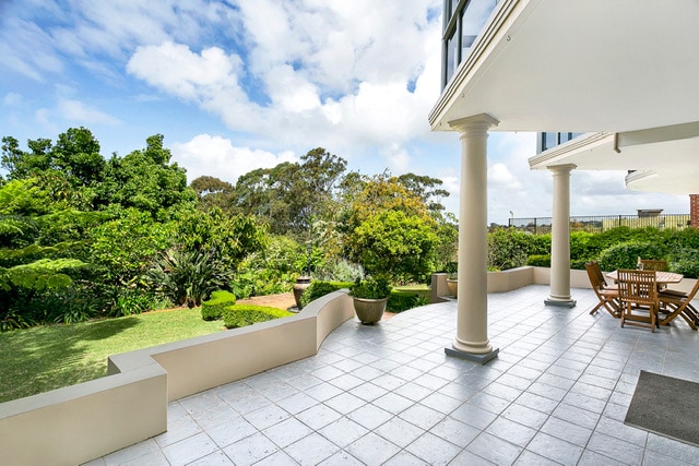 An expansive outdoor patio is showcased, featuring a tiled surface and surrounded by lush greenery and well-maintained gardens. A wooden dining set is positioned nearby, inviting meals in a tranquil setting. The sky above is clear with soft clouds, enhancing the serene environment.