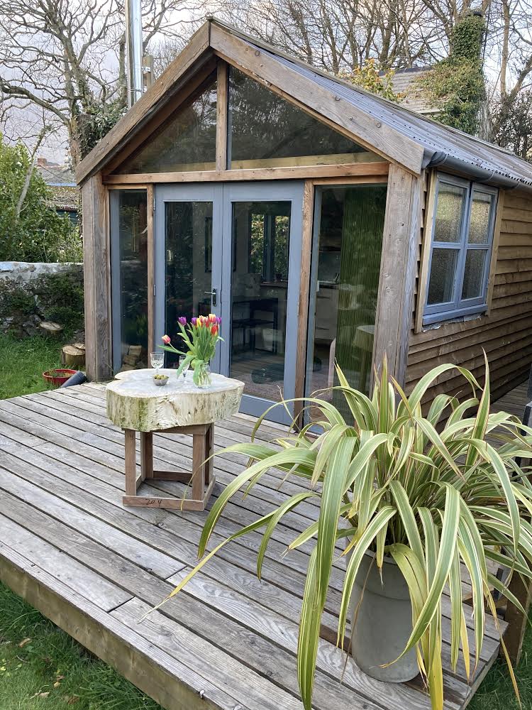 A rustic cabin is shown with a sloped roof and large glass doors, inviting natural light. A circular wooden table with a floral arrangement sits on a wooden deck, surrounded by lush greenery and potted plants. The entrance leads into a cozy interior space.