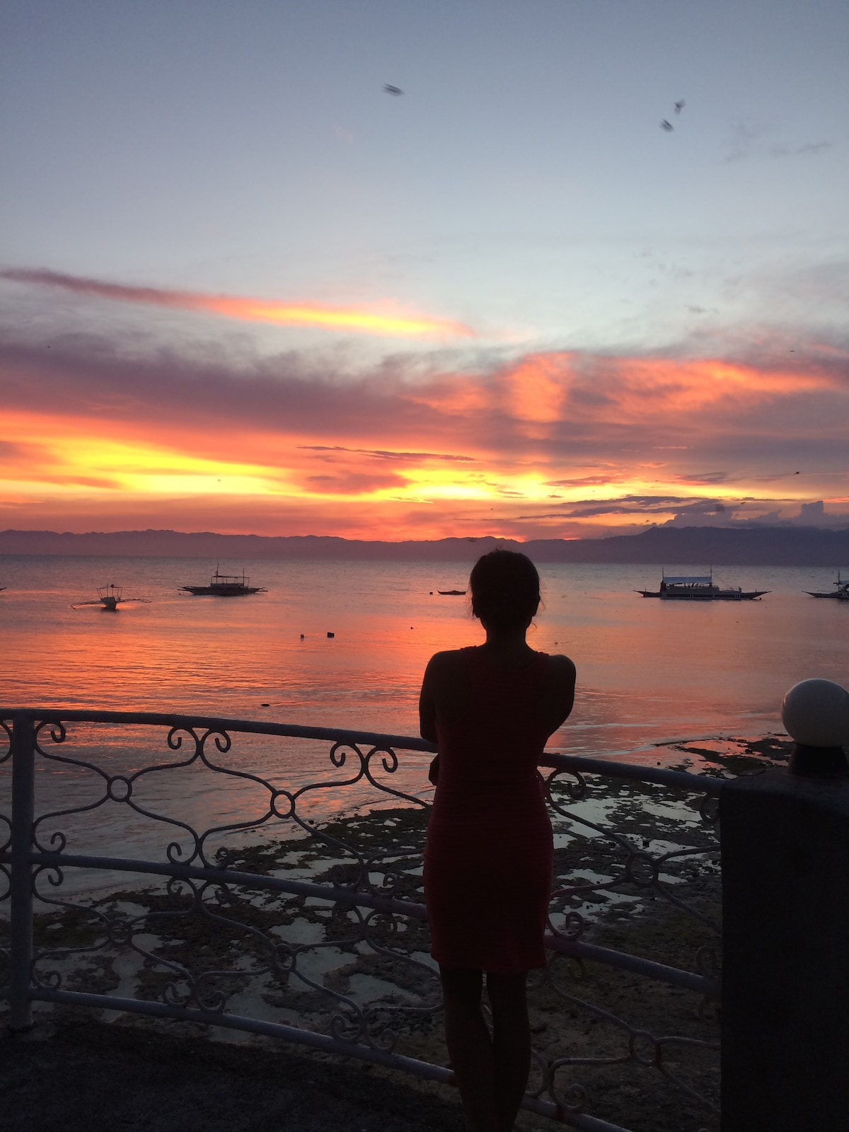 A silhouette of a person stands on a balcony overlooking a serene seascape. The vibrant sunset paints the sky in hues of orange and pink, reflecting on the calm waters. Small boats are visible in the distance, adding to the tranquil atmosphere.