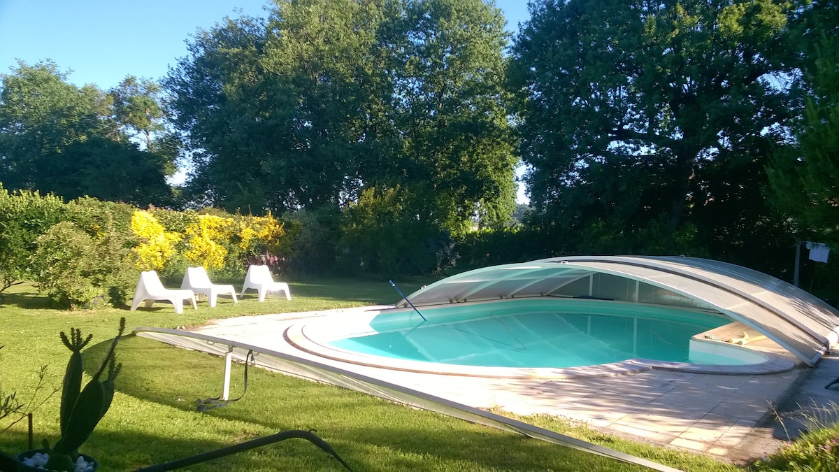 A covered swimming pool is surrounded by a spacious grassy area, with colorful foliage offering a vibrant backdrop. Lounge chairs are positioned nearby, providing a relaxing space to enjoy the outdoors. The tranquil environment is complemented by mature trees, enhancing the peaceful setting.