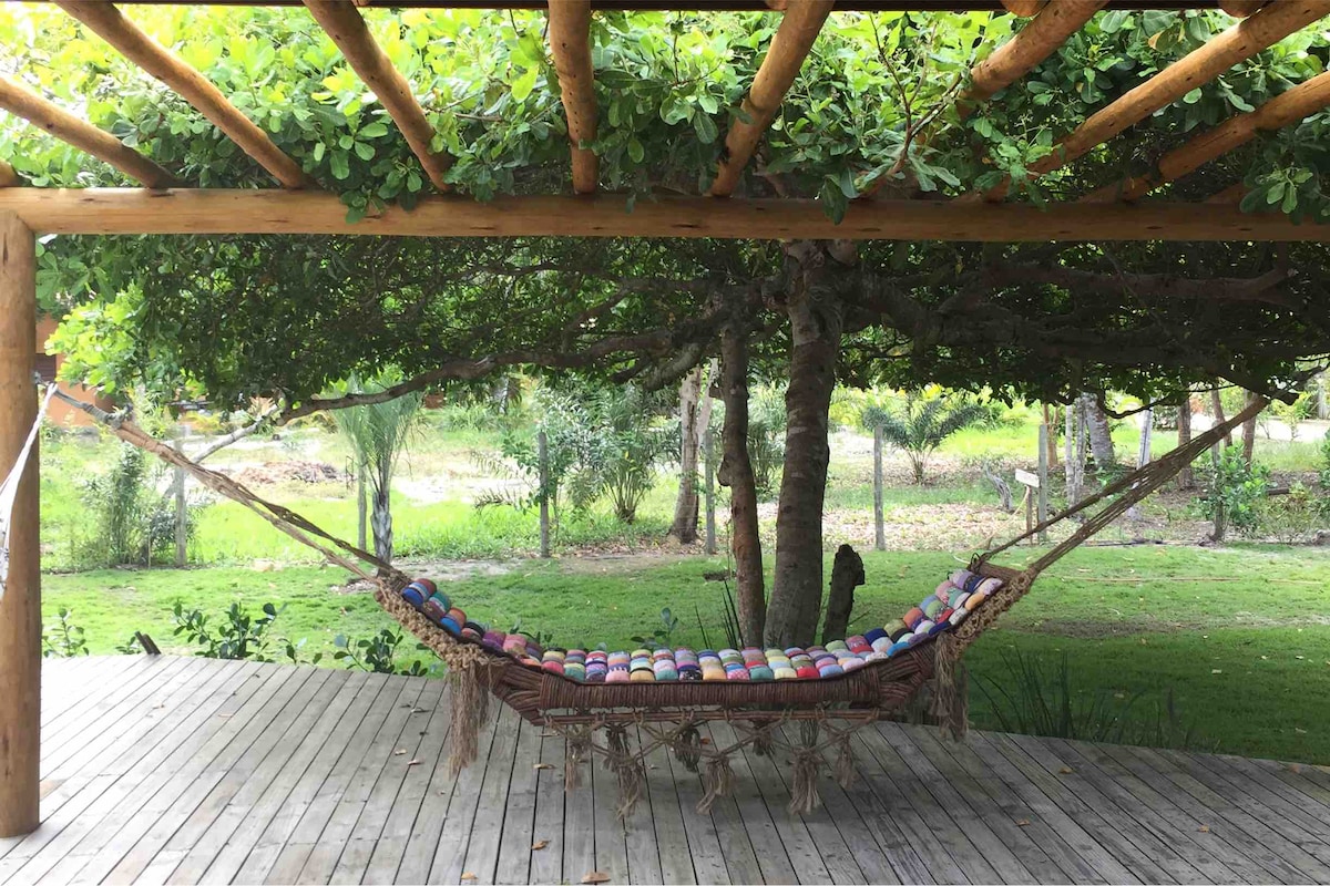 A hammock is gently hung beneath a wooden pergola, surrounded by lush greenery. The deck features wooden flooring, extending towards a grassy area with trees in the background, providing a serene setting for relaxation.