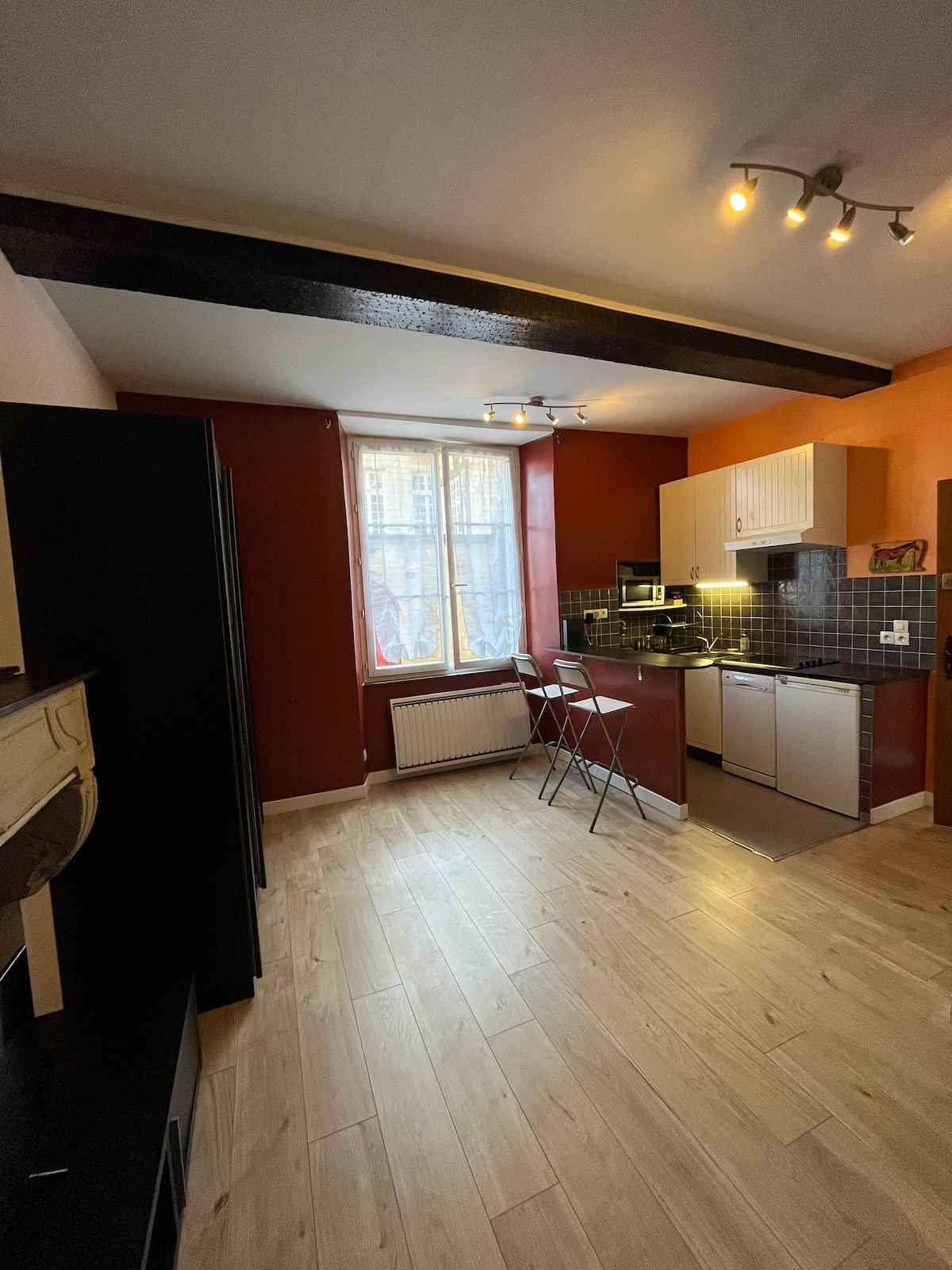 A kitchen area features warm-colored walls and a combination of modern and traditional elements. Counter space is provided alongside a stove, microwave, and refrigerator. Two tall stools are positioned near a window, which offers natural light while showcasing the wooden beams in the ceiling.