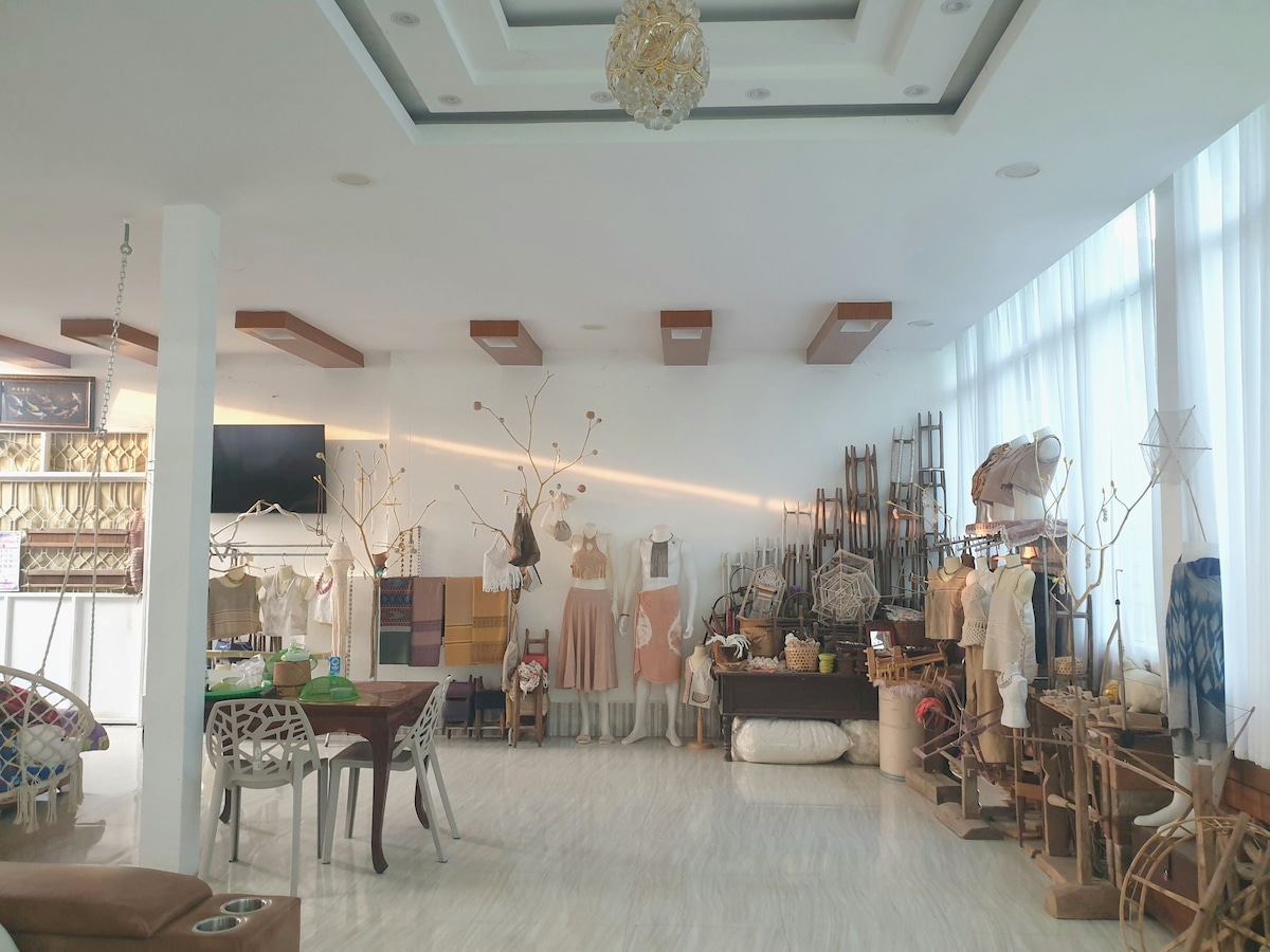 A spacious craft room is filled with various handmade garments displayed on wooden racks. Natural light floods the area through large windows, illuminating the white walls and floor. Tables and chairs are arranged for workspaces, surrounded by artistic decor and craft materials.