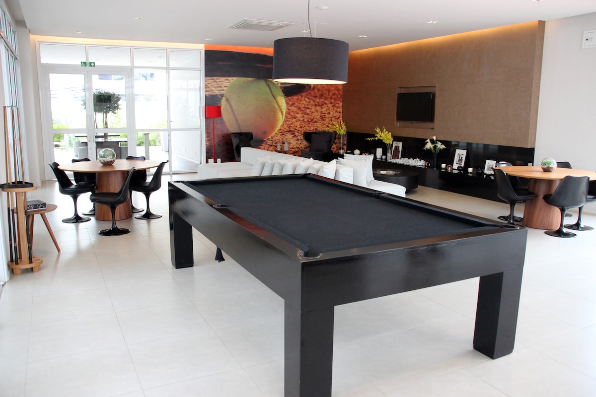 A spacious lounge area is featured, with a large black pool table positioned in the center. Surrounding seating includes stylish chairs and tables. A wall adorned with a vibrant mural adds character, while large windows invite natural light into the inviting space.