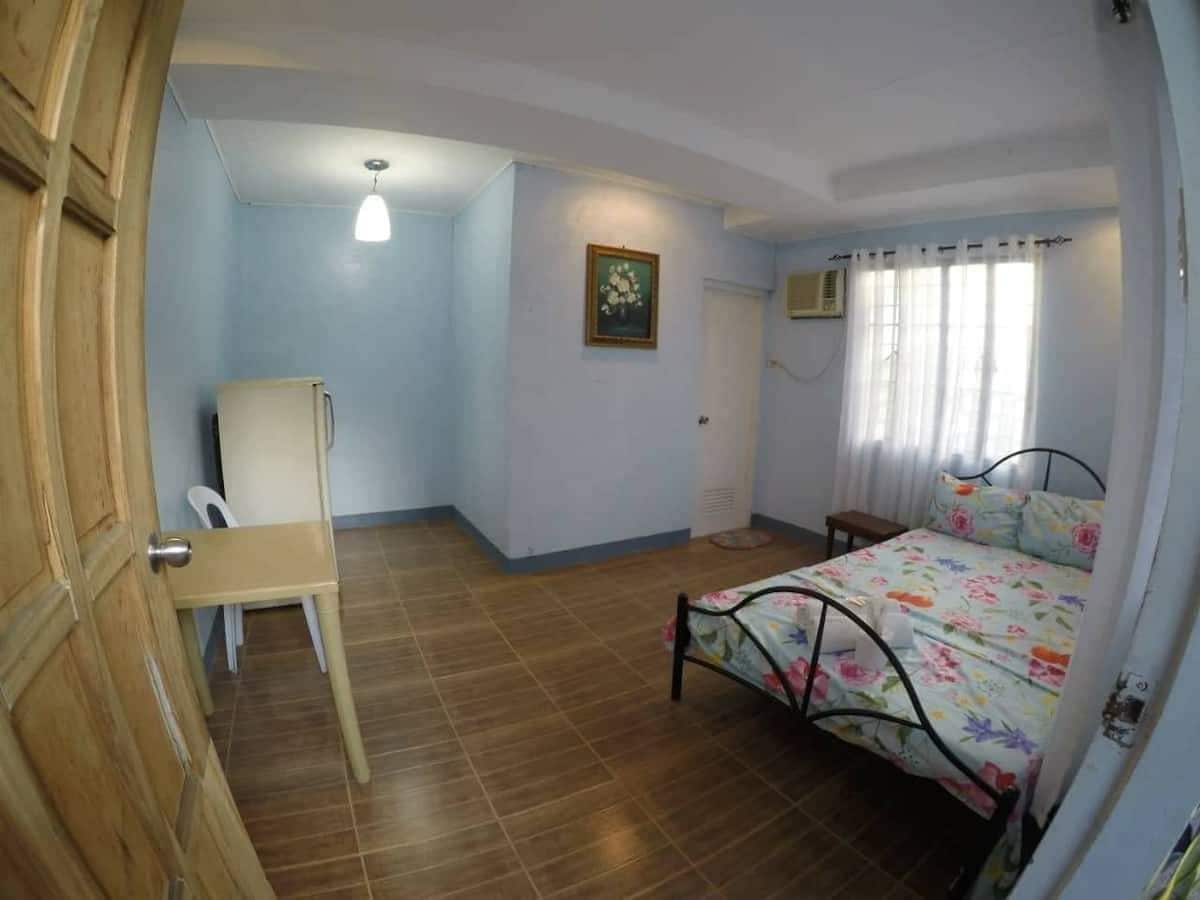 A well-lit room features a floral-patterned bed and a small wooden table with a chair. A refrigerator stands next to the bed, while a window with sheer curtains allows natural light to fill the space. A painting adds a touch of color to the wall.