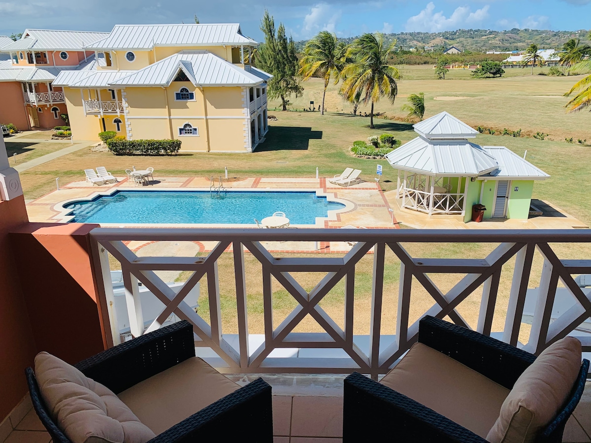 A balcony is shown with two wicker chairs, offering a view of a shared pool surrounded by palm trees. In the background, the landscape extends towards a lush green area, revealing the rhythm of the gentle hills under a clear blue sky.