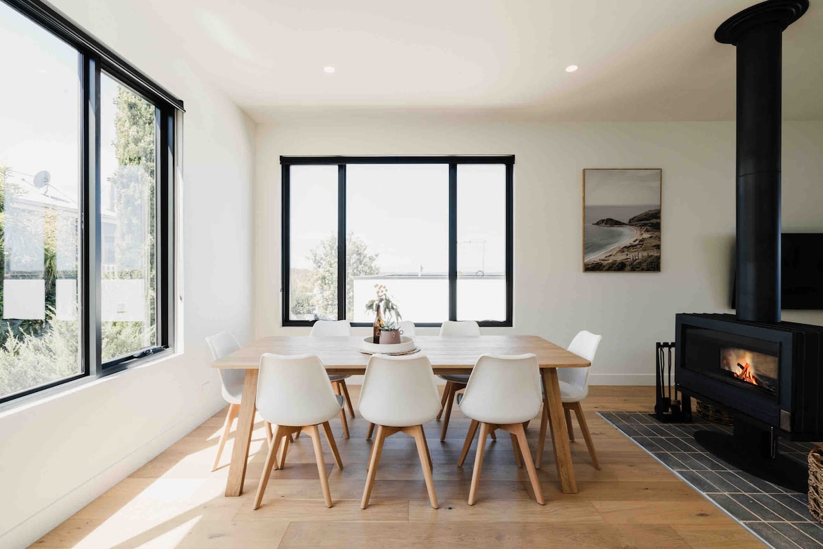 A spacious dining area features a large wooden table surrounded by white chairs, illuminated by natural light from expansive windows. A modern woodburning fireplace stands in one corner, adding warmth to the room, while a scenic artwork is displayed on the adjacent wall.