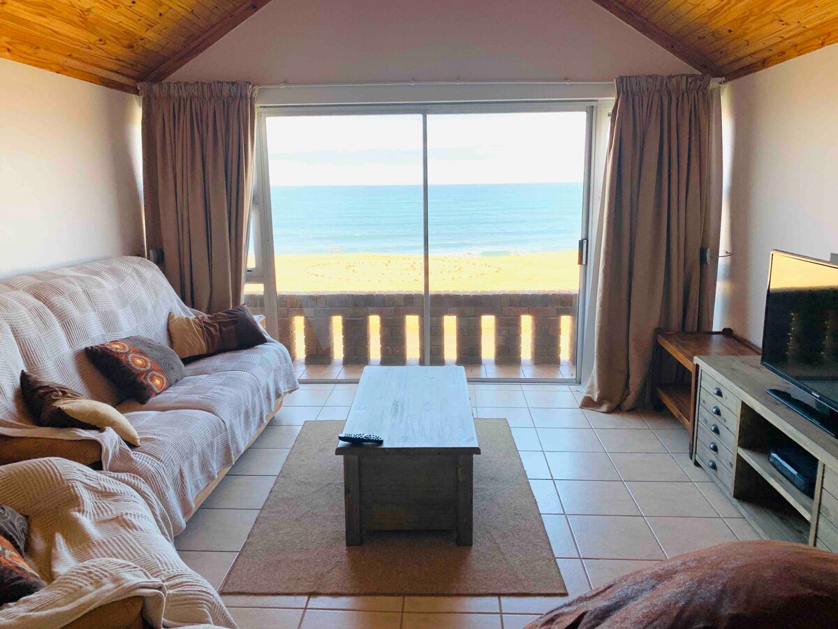 A comfortable living area features a large window overlooking the ocean, allowing natural light to fill the space. A plush sofa is positioned with colorful cushions, accompanied by a wooden coffee table. A flat-screen TV and warm wooden accents enhance the inviting atmosphere.