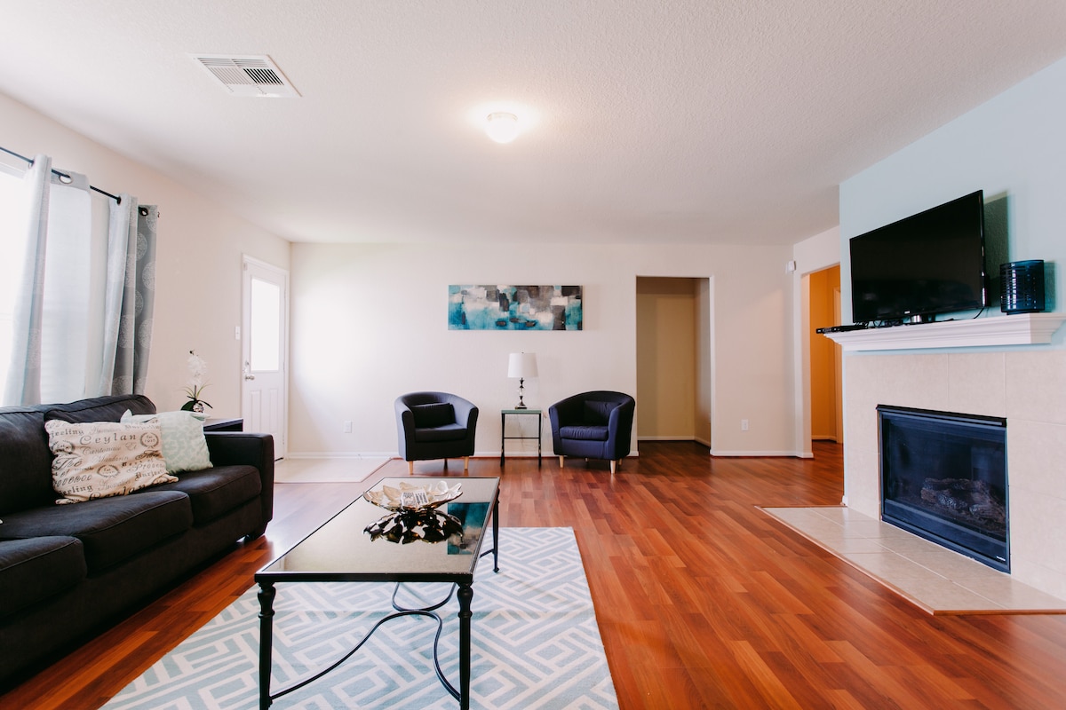 A spacious living room features a dark sofa and two accent chairs positioned around a coffee table. A television is mounted above a tiled fireplace. Natural light filters through the windows, illuminating the wood laminate flooring.