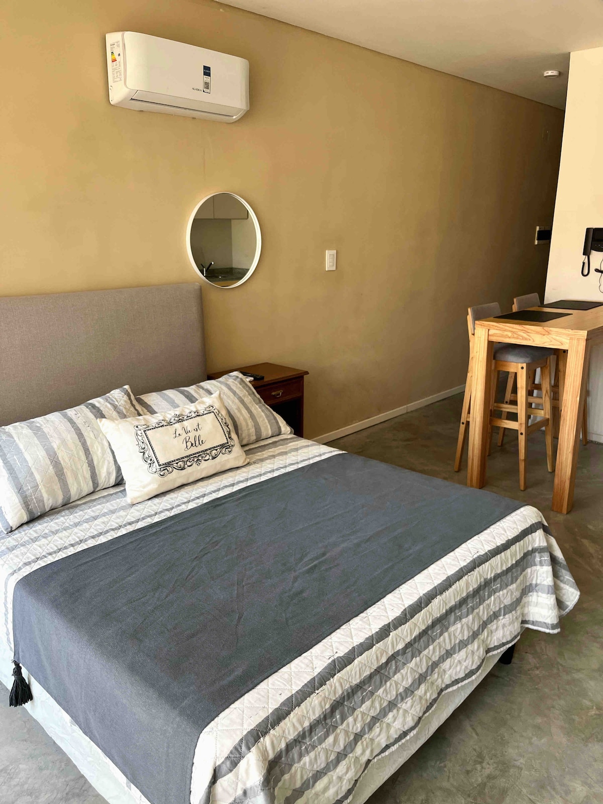 A modern studio features a spacious bed with a textured gray and white blanket. A small round mirror hangs above a bedside table, while a breakfast bar with stools is visible in the background. Air conditioning is mounted on the wall for comfort.