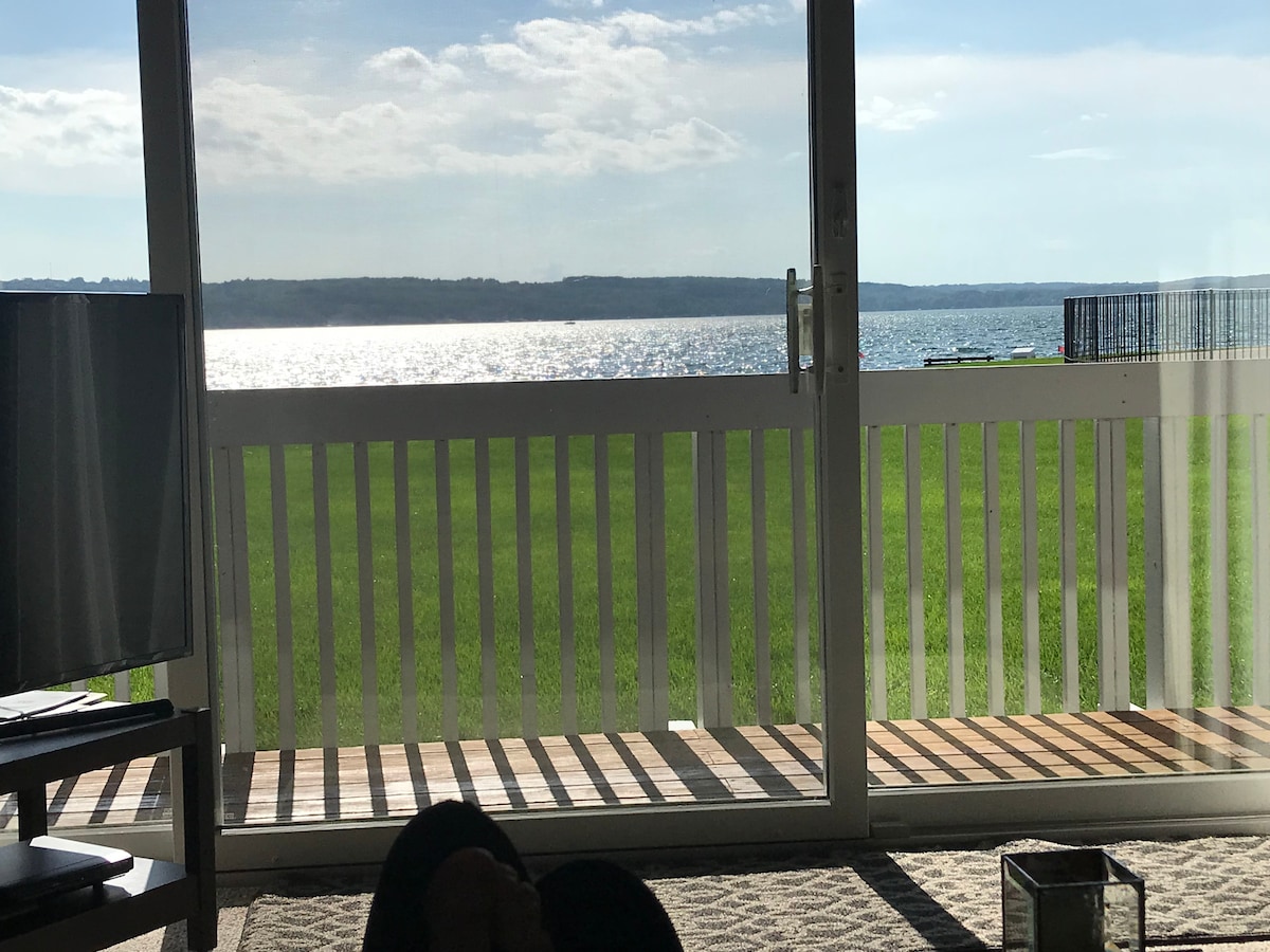 A serene view is captured through large glass doors, revealing a tranquil lake that reflects the sky's light. The grassy shoreline enhances the scenery, while shadows from the slatted railing create a pattern on the floor, inviting relaxation in the space.