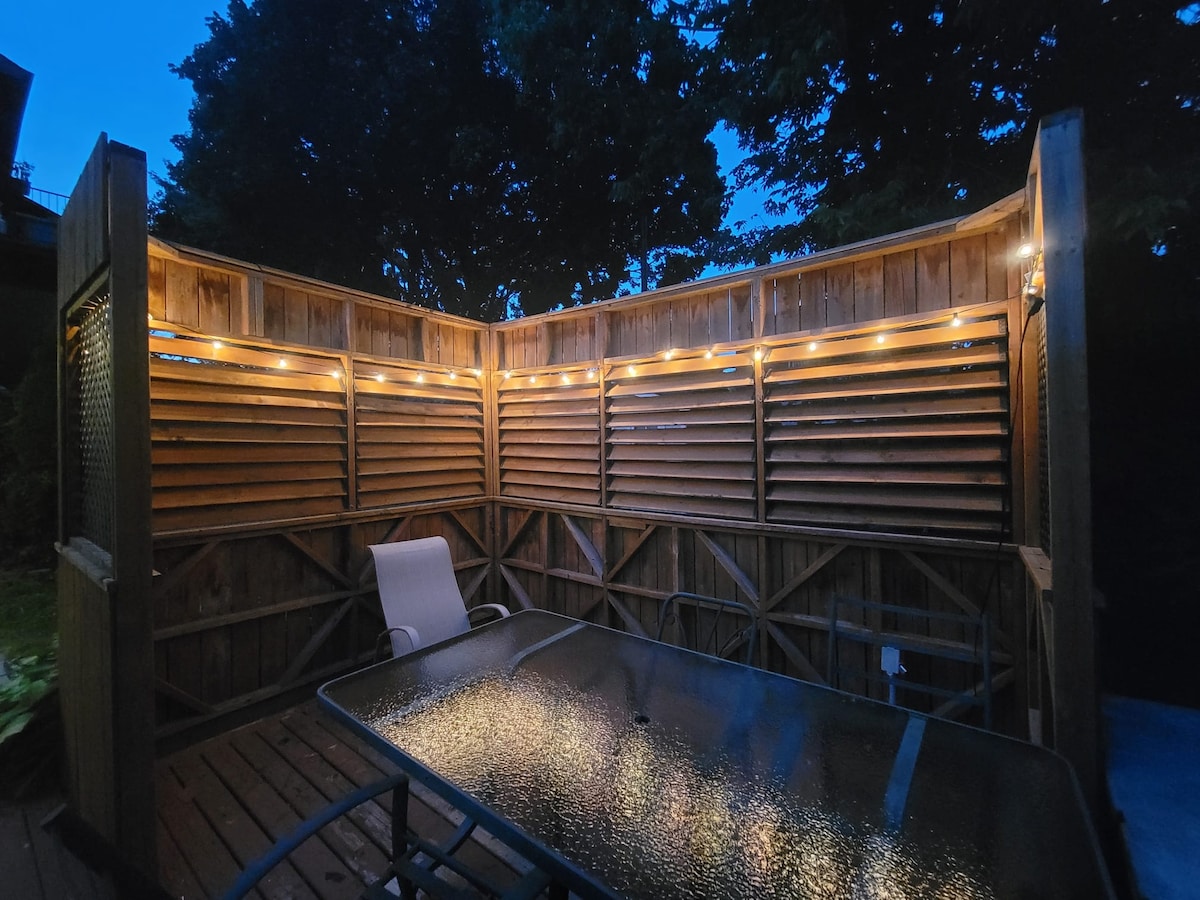 A private outdoor seating area features a glass-top table surrounded by metal chairs. Soft string lights illuminate the wooden slatted walls, creating a cozy atmosphere at dusk. Lush greenery is visible in the background, enhancing the serene environment.