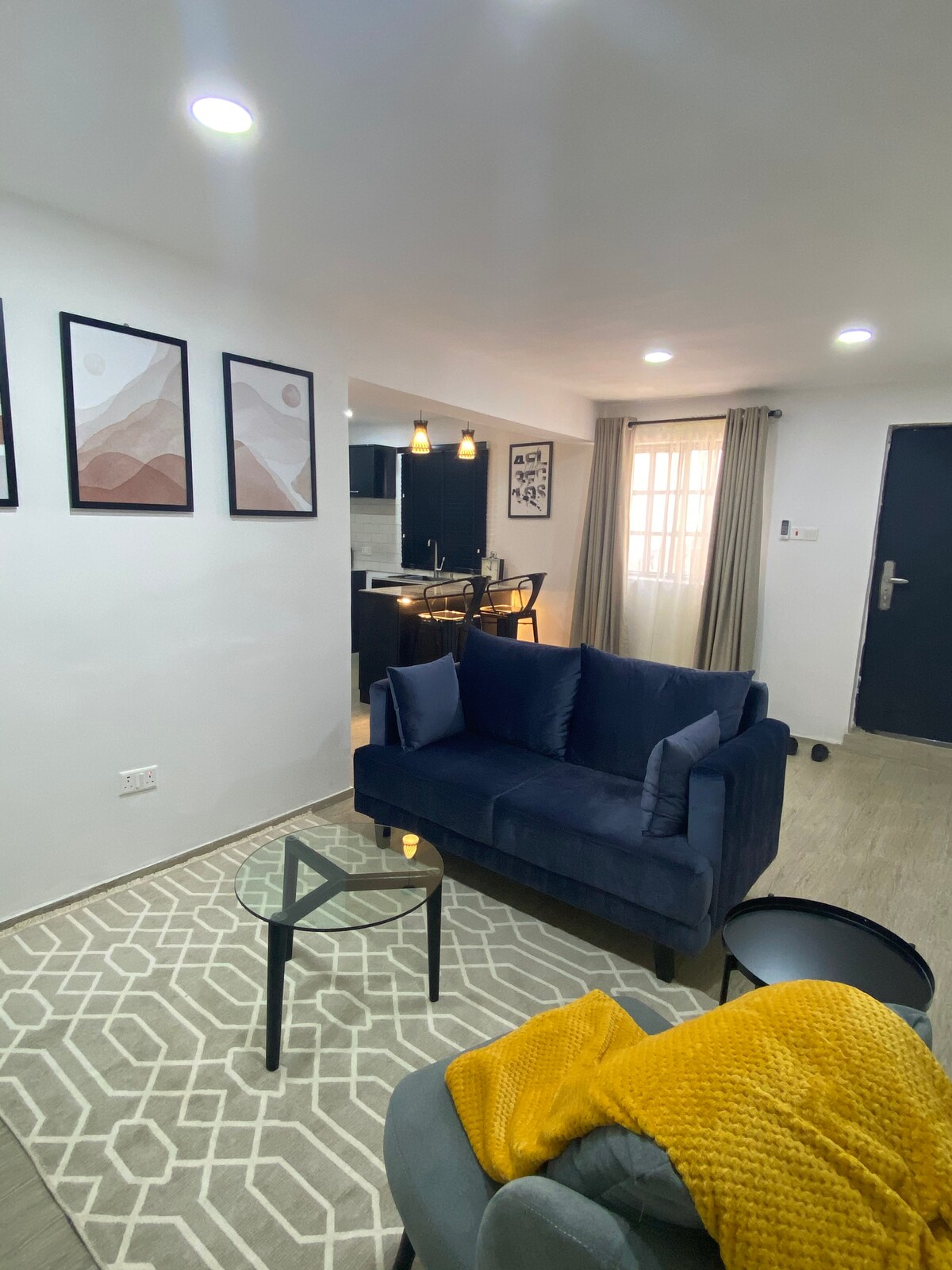 A cozy living area is featured with two comfortable couches in varying styles, complemented by a small glass table. Artwork adorns the walls, and soft light is provided by ceiling fixtures. A glimpse into the kitchen is visible through an open doorway, enhancing the welcoming atmosphere.