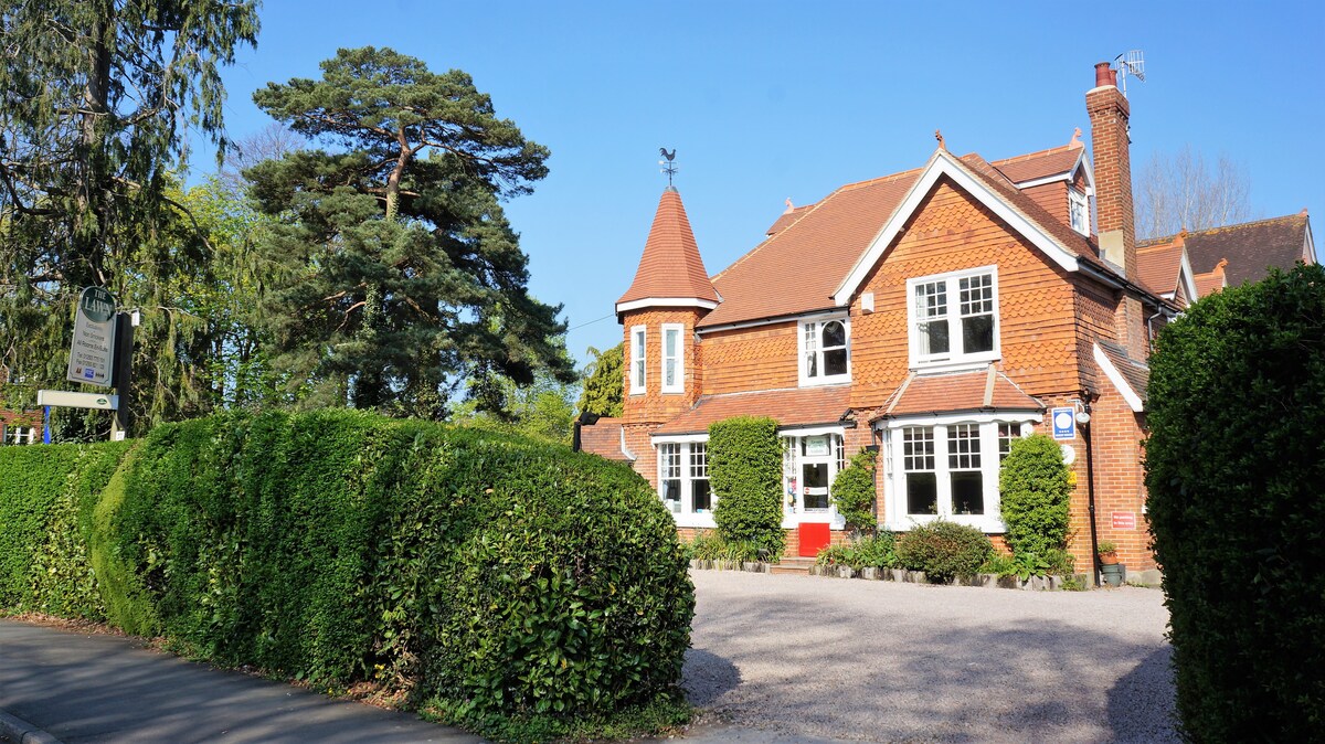 The exterior of a charming red brick guest house is showcased, featuring a distinct turret and a sloped roof. Lush green hedges frame the front, and a gravel driveway leads up to the entrance. Large windows allow natural light to fill the inviting space.