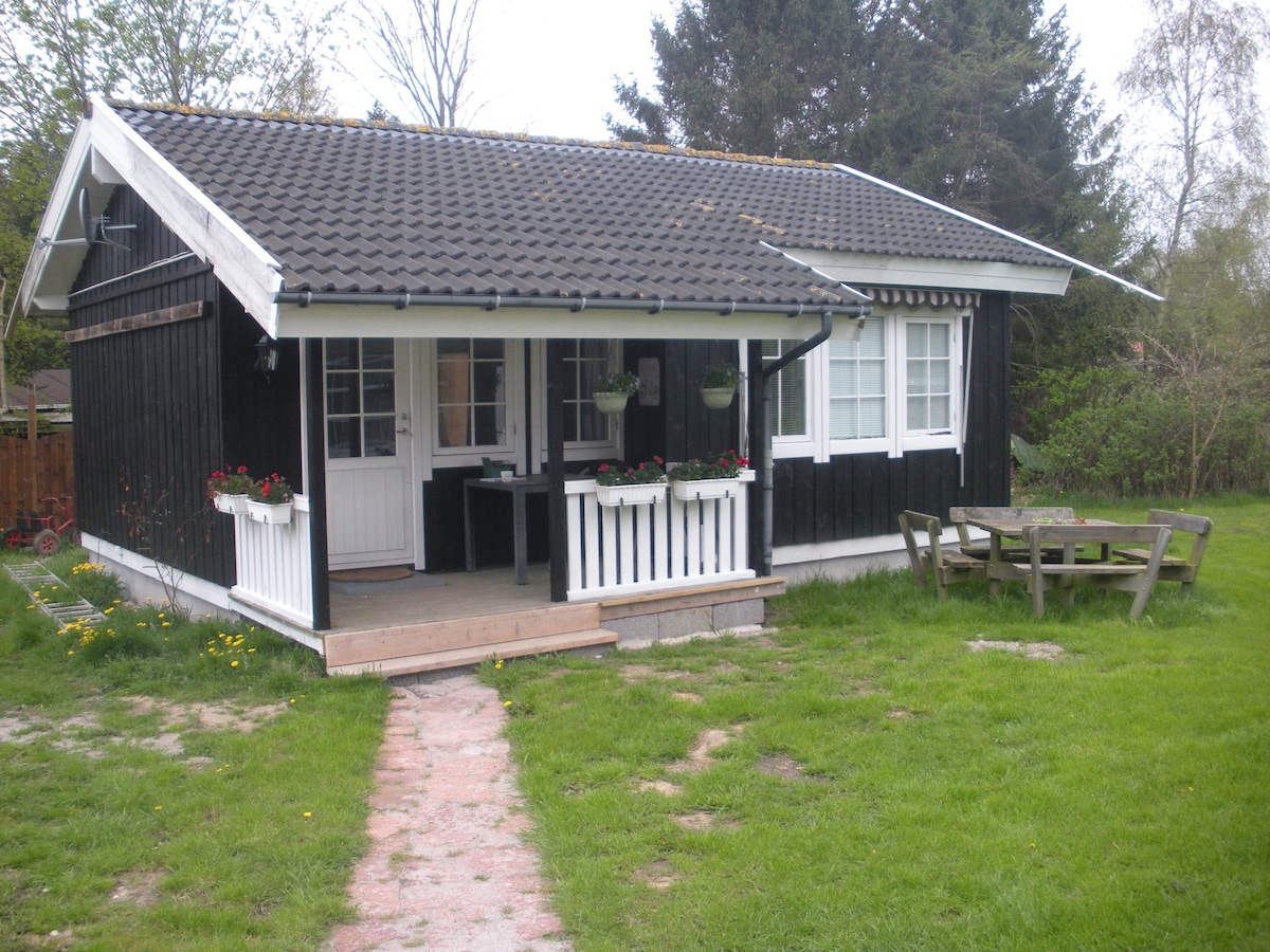 A quaint black wooden guesthouse features a welcoming porch adorned with white railings and potted plants. A wooden table and benches are positioned in the front yard, surrounded by lush green grass and trees, creating a peaceful outdoor space.