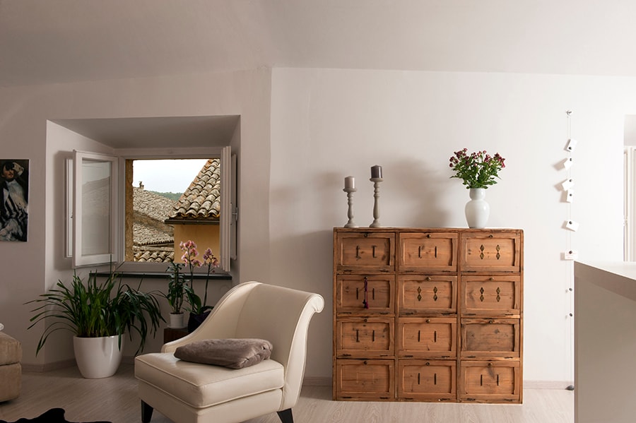 Natural light fills the room, highlighting a wooden storage cabinet adorned with flowers and candles. A soft armchair is positioned nearby, paired with a decorative cushion. An open window reveals rooftops, while potted plants add a touch of greenery to the space.