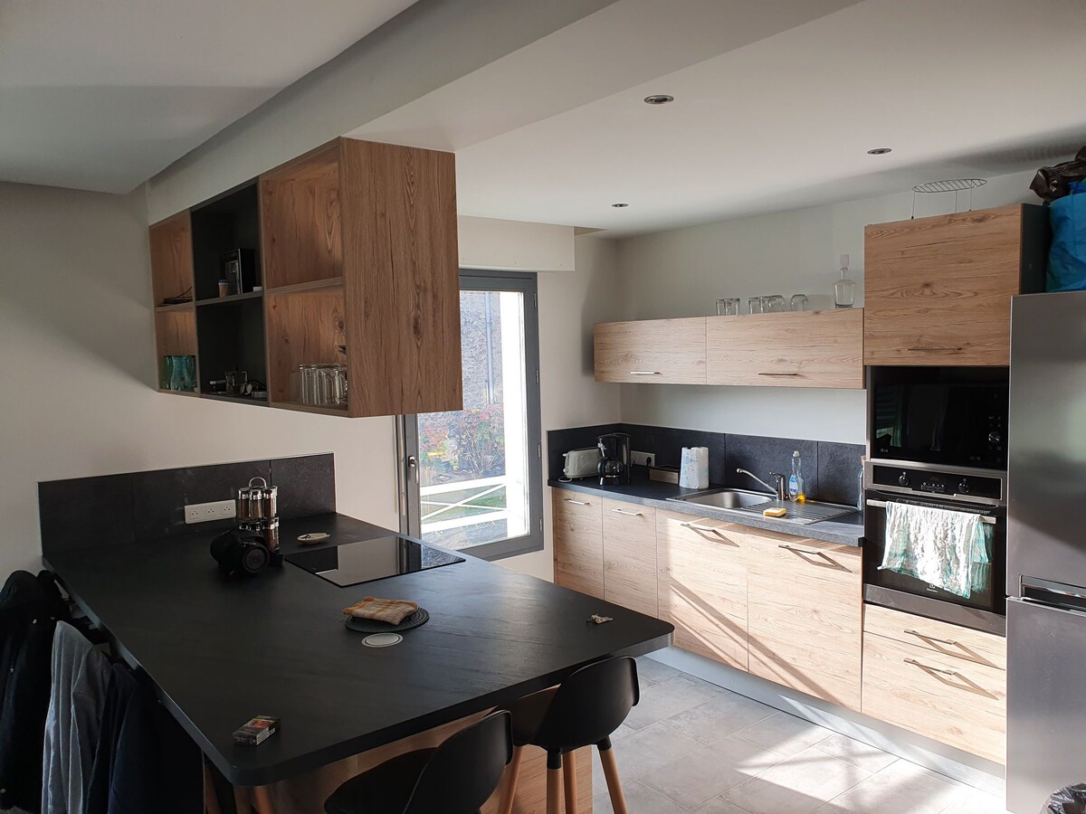 The kitchen features light wood cabinetry and a dark countertop. A large window offers natural light, illuminating the space. An island with bar stools is present, providing additional seating. Kitchen appliances, including an oven and a coffee maker, are integrated into the design.