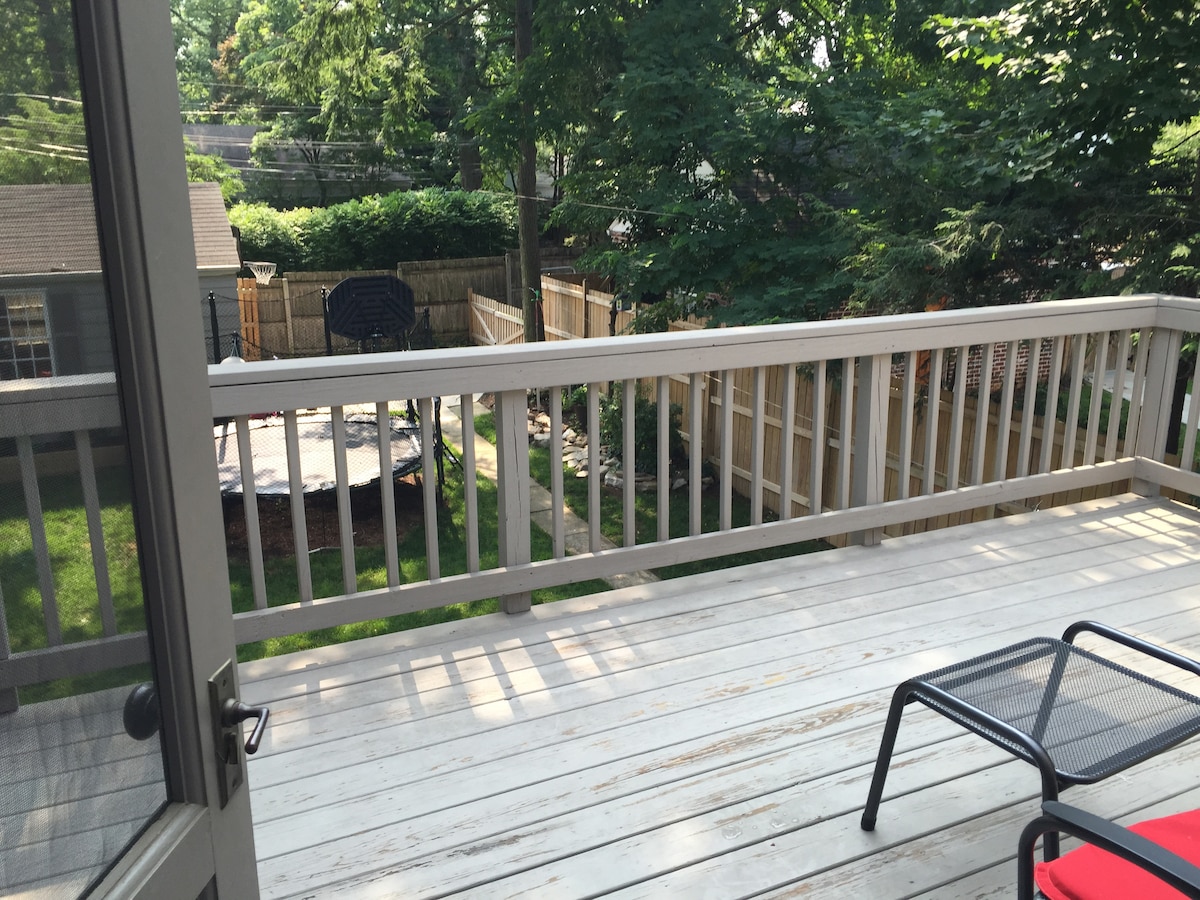 A spacious deck is visible, featuring wooden railings and a view of a landscaped backyard. A basketball hoop is positioned in the yard, surrounded by trees. Lush greenery adds to the calming atmosphere of the outdoor space.